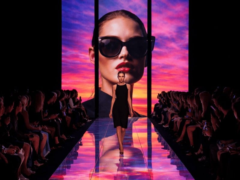 A high-end fashion show featuring an interactive rental LED floor runway and segmented vertical LED video walls. The screens display vibrant, high-contrast fashion visuals that reflect on the catwalk, creating an immersive environment for a brand launch.